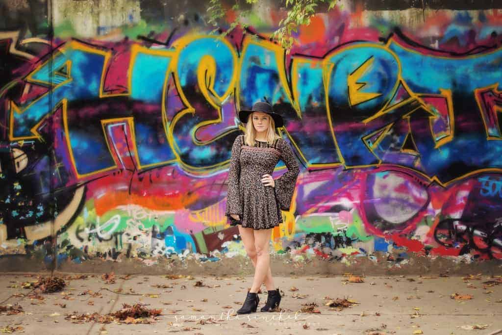 High school senior wearing black hat and stands in front of graffiti wall