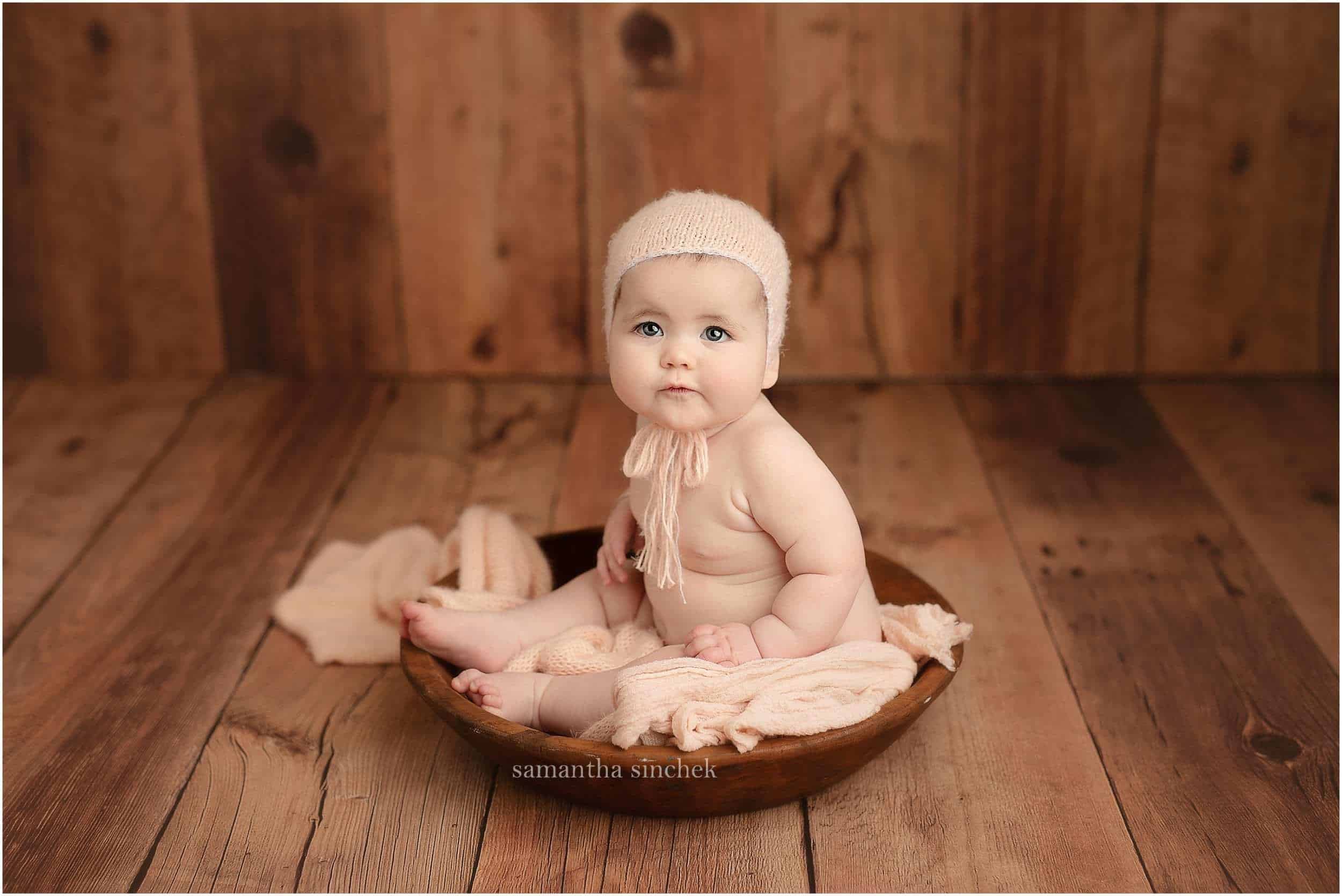 little girl sits in wood bowl wearing pink bonnet at photo session with Samantha Sinchek photography
